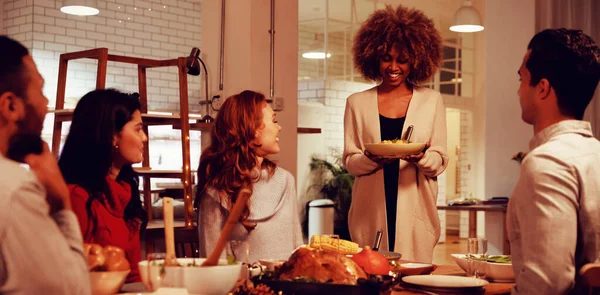 Front view of a young mixed race woman standing and holding a plate of food to serve her four young adult multi-ethnic male and female friends sitting around a table at home for Thanksgiving dinner