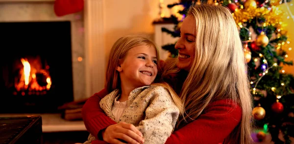 若い白人女性が 飾られたクリスマスツリーの横に座って お互いに微笑みながら クリスマスの時に居間で彼女の膝に彼女の若い娘を抱きしめるの側面図 — ストック写真