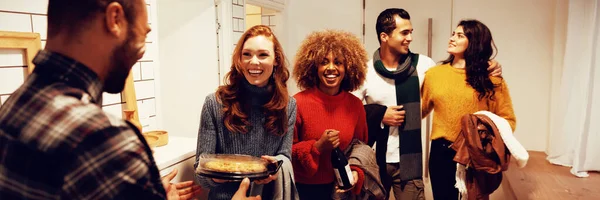 Front view of a group of young adult multi-ethnic male and female friends arriving at a party standing in the hallway of an apartment, one woman carrying a dish of food for the party