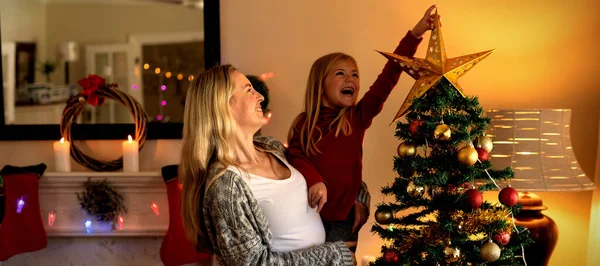 一个快乐的年轻白种妇女抱着她微笑的小女儿 在圣诞节的时候在他们的客厅里装饰圣诞树的正面 — 图库照片