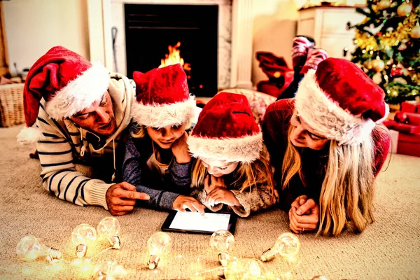 圣诞节期间 一对白种人夫妇头戴圣诞帽 一起使用平板电脑 躺在地板上 他们的小儿子和女儿都在客厅里 — 图库照片