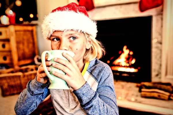 クリスマスの時間に彼のシッティングルームでカップと飲酒を保持サンタの帽子を身に着けている若い白人の少年のフロントビュー — ストック写真