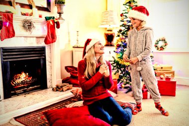 Genç beyaz bir kadının, genç oğlu yanında dururken yerde oturması ön planda. Her ikisi de Noel 'de oturma odasında Noel Baba şapkası takıyor, gülümsüyor ve konuşuyor.