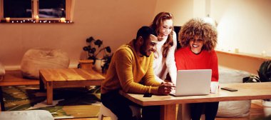 Genç bir karışık ırk adam ve kadın arkalarında duran genç bir Kafkas kadın ile bir masada oturan ön görünümü, birlikte bir dizüstü bilgisayar bakarak ve gece bir dairenin oturma odasında gülümseyerek