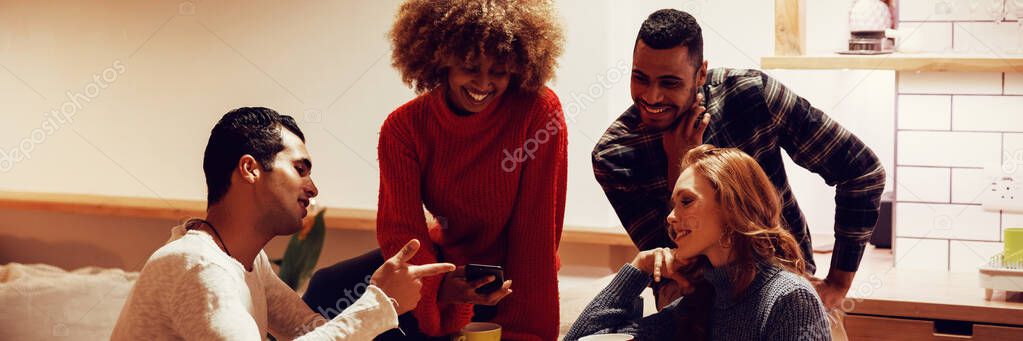 Front view of a group of young adult multi-ethnic male and female friends socialising sitting in the kitchen of an apartment, looking at a phone and smiling