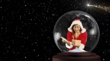 Noel Baba kostümü giyen mutlu, genç beyaz bir kadının kar küresinde bir hediye açması ve siyah bir zemine kar yağması.