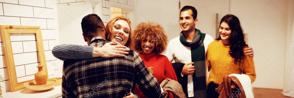 Front view of a group of young adult multi-ethnic male and female friends arriving at a party standing in the hallway of an apartment, one woman carrying a dish of food for the party