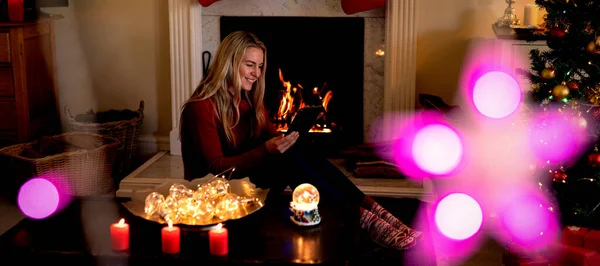 圣诞节时 一位年轻的白种妇女使用平板电脑坐在客厅的壁炉前 前景上挂着圣诞明星装饰 — 图库照片