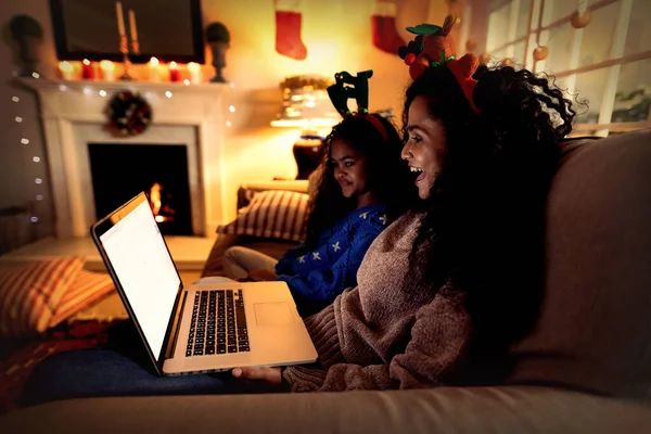 クリスマスに彼女の若い娘とソファに座って ノートパソコンの笑顔とお祝いの帽子を身に着けている混合レースの女性の側面図 — ストック写真