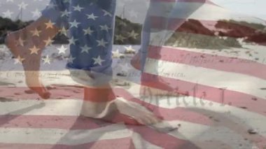Amerikan bayrağının ABD Anayasası metniyle dalgalanması kumsalda çıplak ayakla yürüyen bir çiftin üzerinden geçiyor..
