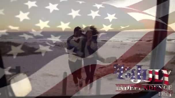 混合されたレースのカップルがビーチを歩く上で独立記念日のテキストを振っている米国の国旗のアニメーション 米国の国旗と休日のコンセプトデジタル組成物 — ストック動画