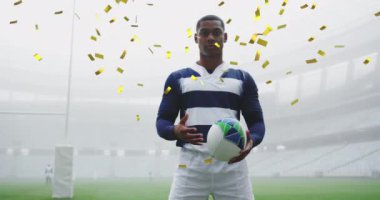 Altın konfeti animasyonu karışık ırktan erkek rugby oyuncusunun üzerine düşüyor. Kameraya bakıyor, arka planda bir rugby topu tutuyor.