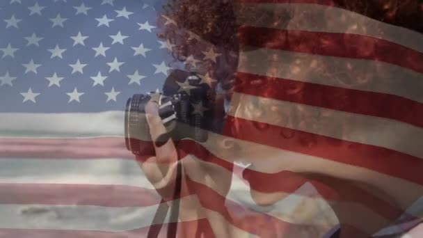 背景是美国国旗飘扬在混血儿身上的动画 背景是夏季度假时 她带着相机在海滨拍照 美国社会多样性概念数字构成 — 图库视频影像