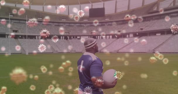 空のスタジアムでラグビーボールを保持ラグビー選手の上に浮かんでいる3Dコロナウイルス細胞のアニメーション グローバルCovid 19コロナウイルスパンデミックスポーツコンセプトデジタル的に生成された画像 — ストック動画