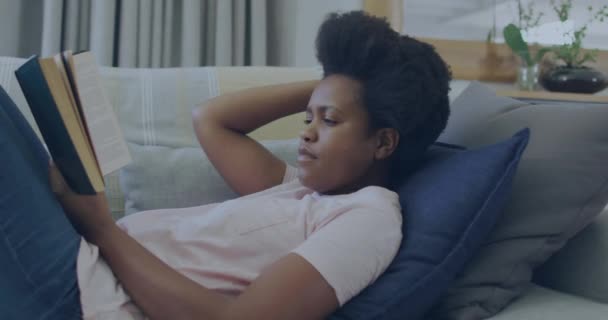 アフリカ系アメリカ人女性が自宅でソファーで本を読んでいるエコアイコンとデータ処理のアニメーション グローバルグリーンエネルギー コネクション コンピューティング データ処理のコンセプトをデジタル生成したビデオ — ストック動画