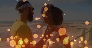 Animation of glowing yellow spots over african american couple embracing each other at the beach. Love and relationship concept