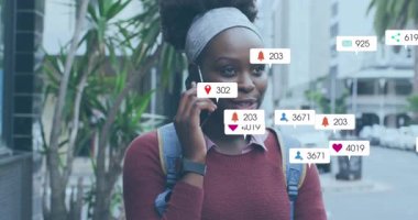 Animation of multiple social media icons over african american woman talking on smartphone outdoors. Social media networking and technology concept