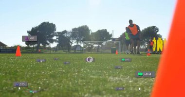 Turuncu önlüklü oyuncu, futbol antrenmanında hoparlörde sinyal karıştırıyor, çimlerin üzerinde hareketli ölçüler yükseliyor. Spor eğitimi, çeviklik, takım çalışması, dijital analitik, atletik performans, açık hava aktivitesi, fitness