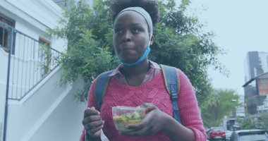 Elinde salata kabı ve çatal, pembe kazaklı kadın merdivenin dışında duruyor, maskeli. Açık hava, şehir, yaşam tarzı, gündelik, modern, sağlıklı, Hillary