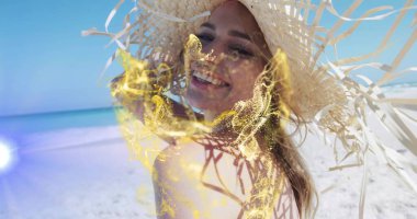 Smiling woman looking back at lens on white sandy beach, with straw hat, swimsuit, golden particles. Tropical, leisure, vacation, summer, glamour, nature, lifestyle