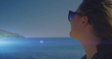 Gazing woman wearing sunglasses, patterned shirt at shore, lens flare crossing scene, copy space. Seascape, adventure, tranquility, outdoor, freedom, landscape, serenity