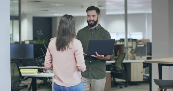 Discussing work two coworkers holding laptop while conversing in office, with desks, plant, stool. Collaboration, teamwork, modern, professional, productivity, communication, workspace