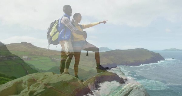 Leaning couple in yellow windbreaker embracing and pointing to sea from cliff, with hiking backpack. Adventure, exploration, landscape, nature, freedom, outdoor, travel