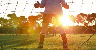 Defending goalkeeper wearing padded gloves and cleats on park grass pitch, with soccer net backdrop. Sports, team, training, fitness, outdoor, activity, youth