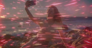 Holding smartphone woman capturing sunset selfie on rocky shore by ocean, red light streak overlay. Landscape, tranquility, leisure, vibrancy, reflection, urban, nature