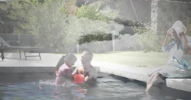 Baba kızını havuzda tutuyor yüzme dersi için tekmeliyor, anne güneş kremi sürüyor. Aile, rahatlama, açık hava, su, boş zaman, güneş ışığı, kaynaşma