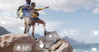 Standing couple with backpacks on jagged rock, pointing toward sea with social icons, copy space. Adventure, exploration, scenic, rugged, outdoor, travel, discovery