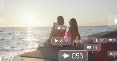 Couple wearing plaid shirt and pink top pointing toward sunset at seaside pier, with social icons. Romance, leisure, relaxation, scenic, communication, technology, lifestyle