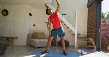 Performing standing side stretch man in red top on blue mat at home, with water bottle. Fitness, wellness, exercise, modern, interior, activity, motivation