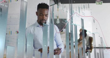 Analyzing business-casual man viewing touchscreen device in open-plan office, with financial charts. Technology, teamwork, innovation, modernity, productivity, corporate, analytics