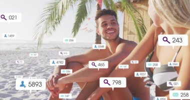 Smiling couple in swimwear sitting under palm tree on sandy beach, with floating social media icons. Tropical, relaxation, communication, leisure, vibrant, outdoor, connection