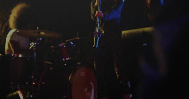 Singer in patterned pants performing on stage holding mic stand, with red drum kit and lights. Performance, concert, nightlife, stage, rhythm, entertainment, acoustic