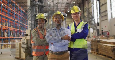 Standing warehouse workers crossing arms in warehouse, with hard hats, safety vests and pallets. Industrial, logistics, safety, teamwork, construction, infrastructure, management
