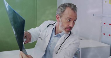 Pointing doctor in lab coat holding radiograph film on wall in exam room, with stethoscope, cabinet. Medical, healthcare, professional, diagnostic, clinical, inspection, diagnostic imaging