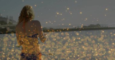 Standing woman wearing blue bikini at sea edge by breakwater, with bokeh light reflections. Seascape, tranquility, sunset, leisure, relaxation, nature, minimalism