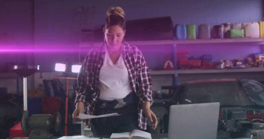 Reviewing papers plaid-shirted mechanic checking manual and laptop on garage bench with grinder. Industrial, craftsmanship, productivity, technological, maintenance, precision, workspace