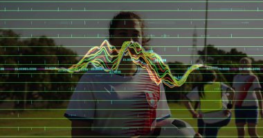 Futbol forması giyen bir futbolcu sahanın ortasında, üzerinde grafiği olan bir top tutuyor. Atletizm, takım çalışması, açık hava, spor, rekabet, coşku, motivasyon