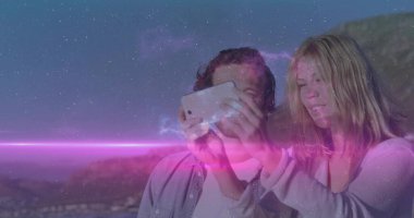 Standing couple holding smartphone and capturing selfie at coastal cliff at dusk, cosmic flare. Romantic, adventure, scenic, twilight, celestial, outdoor, exploration