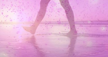 Sahilin altın saatinde, sığ sularda yürüyen kadın bacakları ve ayakları, pembe bokeh örtüsü. Deniz manzarası, sükunet, dinginlik, doğal, şiirsel, yansıtıcı, sıcak tonlar.