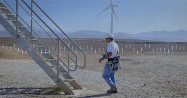 Technician gripping handrail and securing safety lanyard starting climb for wind turbine inspection. Renewable energy, sustainability, industrial, technology, outdoor, engineering, environmental