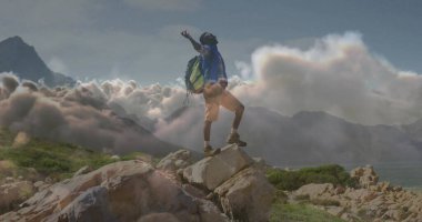 Standing hiker raising arms atop rocky outcrop in mountains, with green backpack and hiking boots. Adventure, exploration, accomplishment, wilderness, outdoor, rugged, triumph