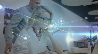 Woman taking office notes while colleague approaches offering tablet and showing network overlay. Collaboration, teamwork, innovation, digital, modern, business, communication