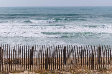 Fransa, Lacanau 'daki sahil boyunca kum tepelerini erozyondan koruyan ahşap bir çit. Arka plandaki okyanus dalgaları doğal dokuları olan sakin bir kıyı manzarası yaratır.