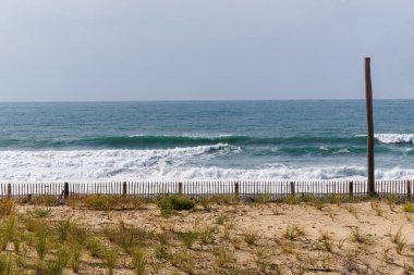 Fransa, Lacanau 'daki sahil boyunca kum tepelerini erozyondan koruyan ahşap bir çit. Arka plandaki okyanus dalgaları doğal dokuları olan sakin bir kıyı manzarası yaratır.