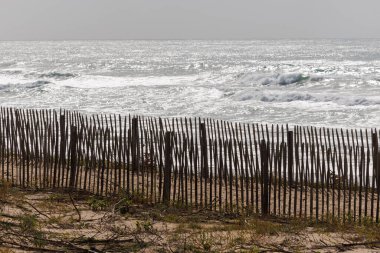 Fransa, Lacanau 'daki sahil boyunca kum tepelerini erozyondan koruyan ahşap bir çit. Arka plandaki okyanus dalgaları doğal dokuları olan sakin bir kıyı manzarası yaratır.