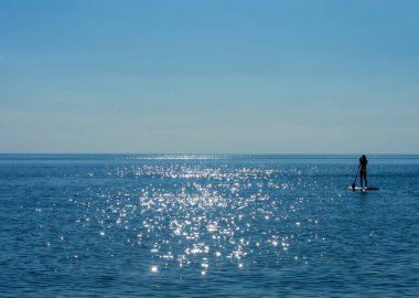 Güneş parlayan bir deniz ve ufukta küreği olan şişme bir tahtanın üzerinde bir kız.